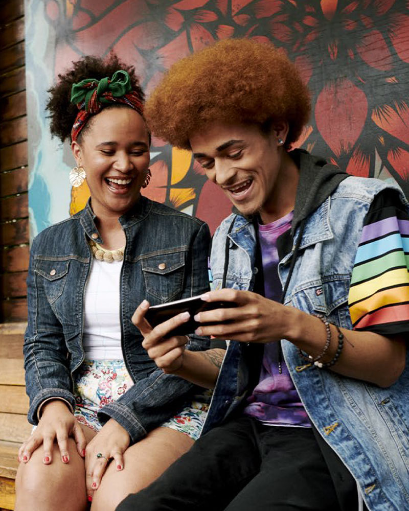 A man and woman are smiling as they watch him play a game on his phone in front of a large colourful mural.